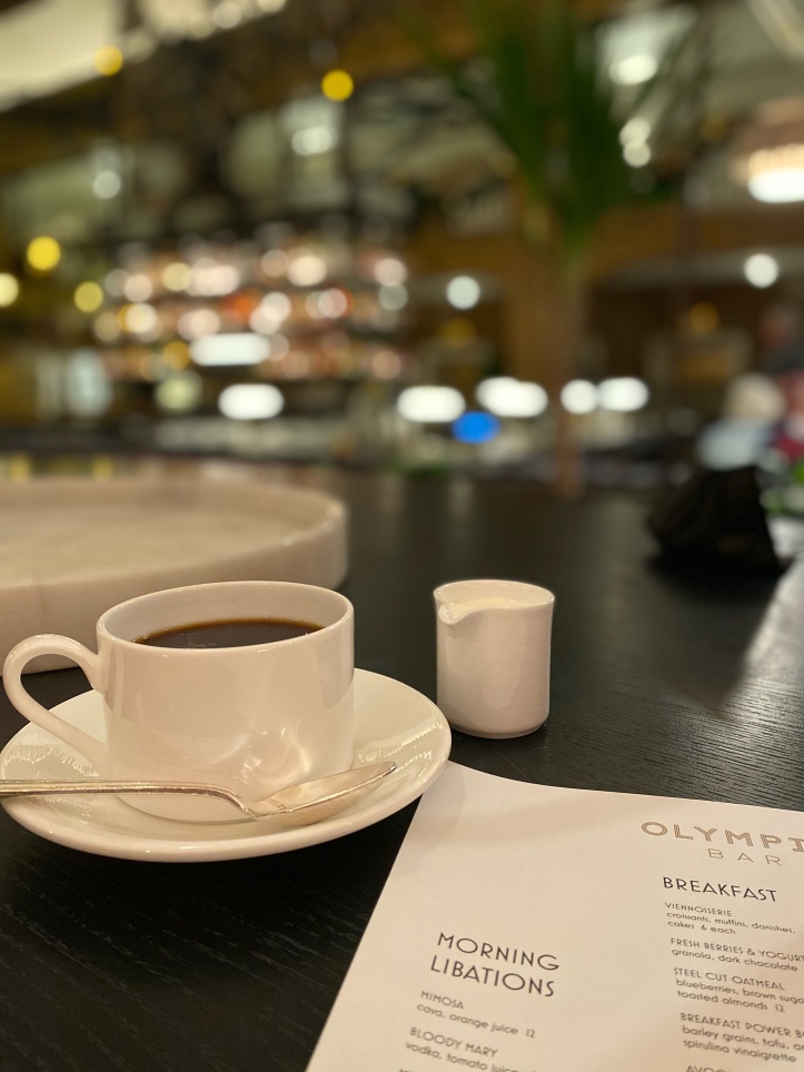 a small white cup of black coffee sitting on top of a white saucer, with a small white pitcher of cream adjacent to it. a white and black font restaurant menu sits in the bottom right corner of the image 